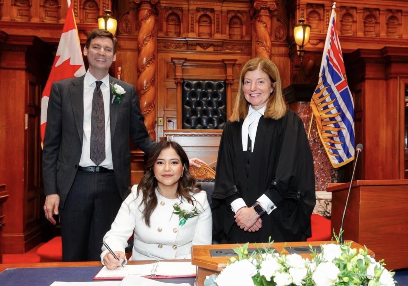 Amna Shah at a desk during a ceremony in the B.C. Legislature, with two people standing beside her.