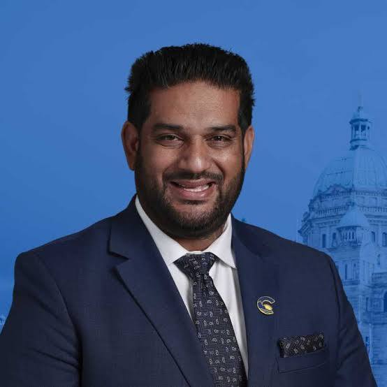 Portrait of Mandeep Dhaliwal in a dark suit against a blue background.